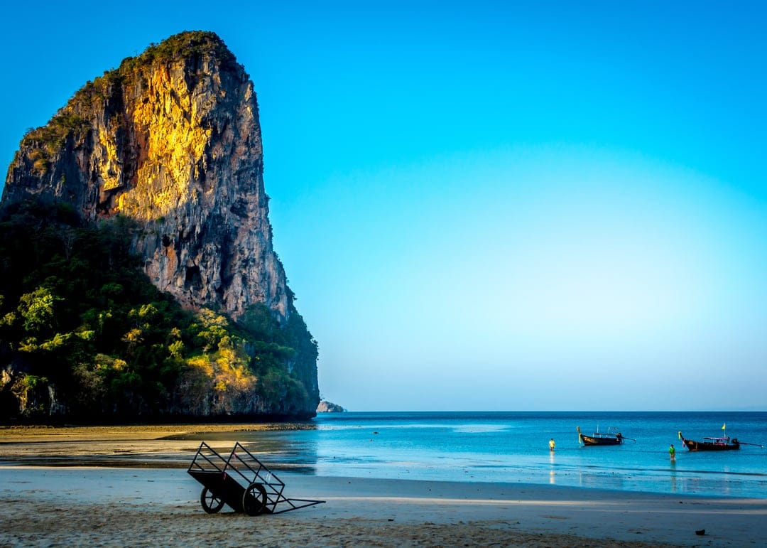 Limestone cliffs tower over traditional longtail boats in turquoise water during a golden tropical beach sunrise.