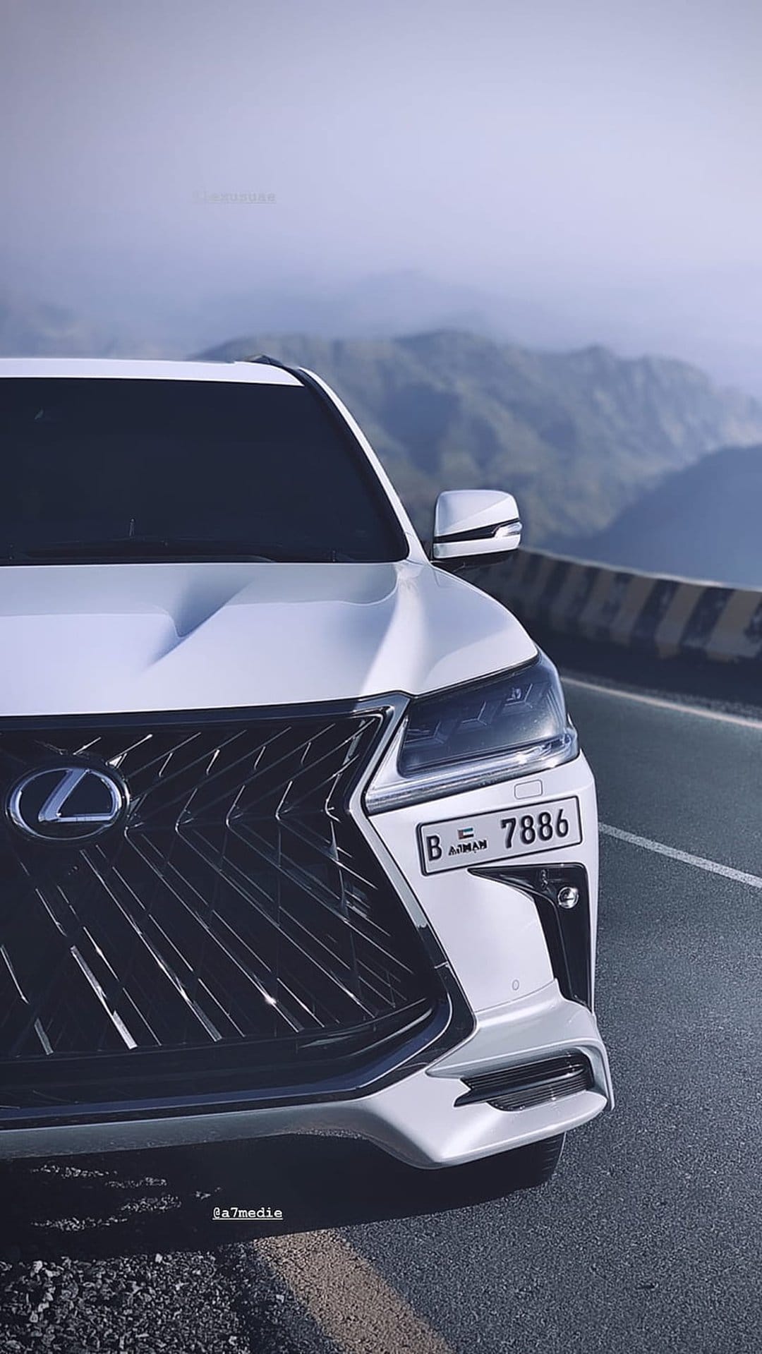 White Lexus SUV with a prominent grille parked on a scenic mountain road overlooking misty peaks and a guardrail.