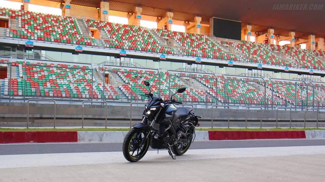 Black Yamaha MT-15 motorcycle parked on a grey racetrack in front of red, green, and white stadium seats.