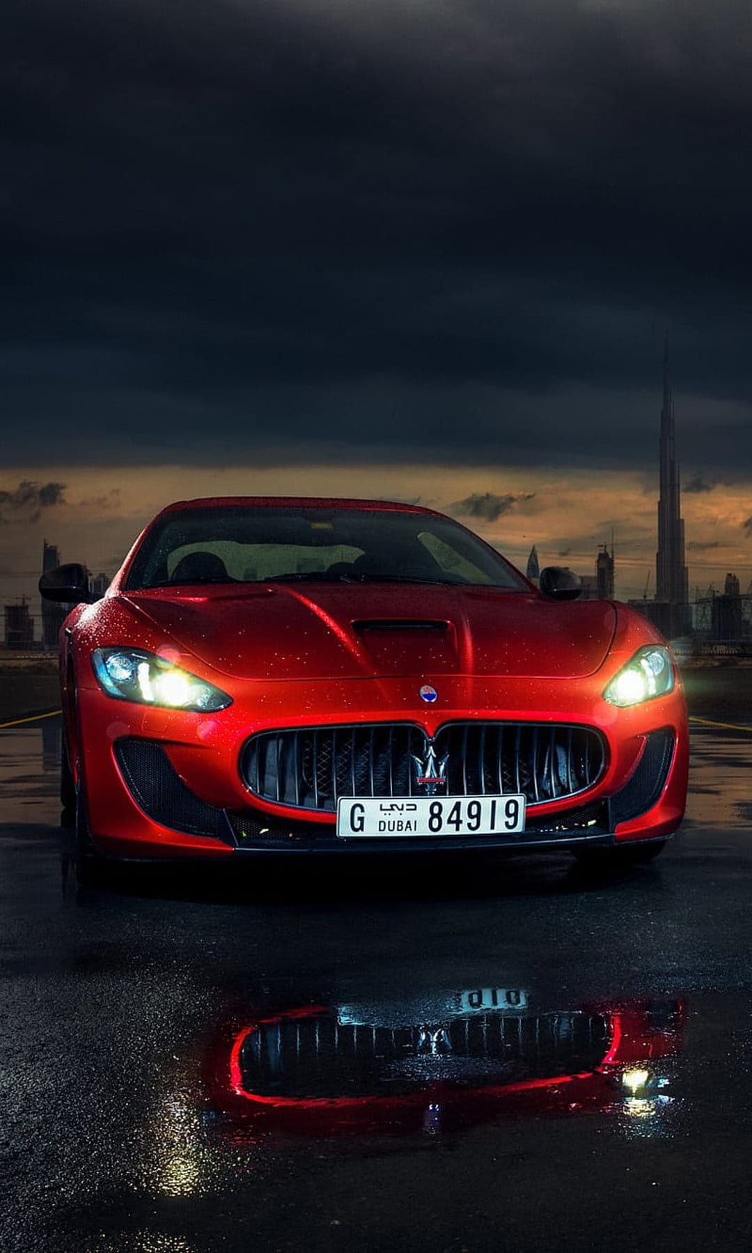 Red Maserati GranTurismo on a wet road with the Burj Khalifa under a cloudy sky during a Dubai evening.