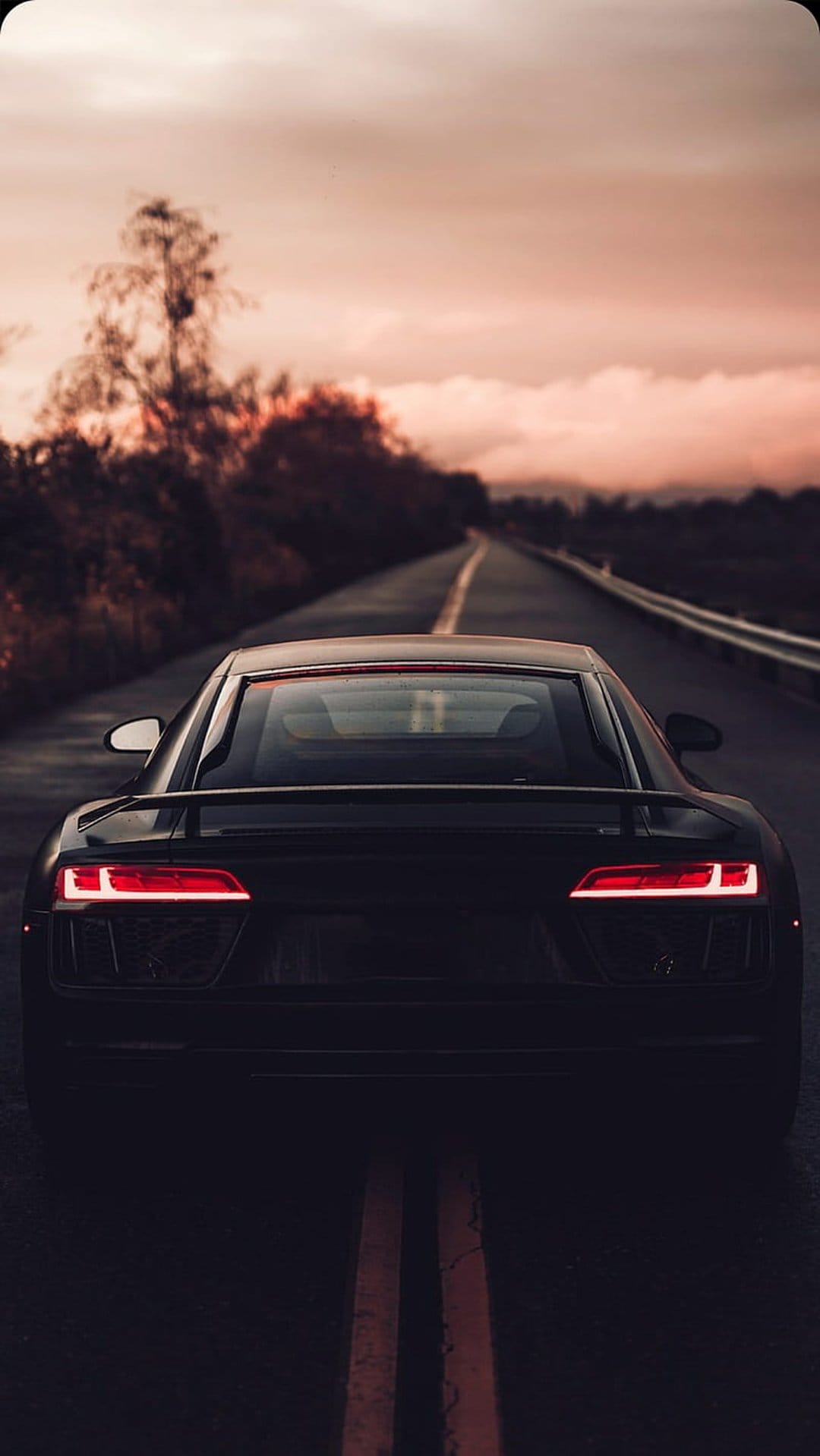 Black Audi R8 sports car with glowing red taillights driving on a mountain road during a vibrant orange sunset.