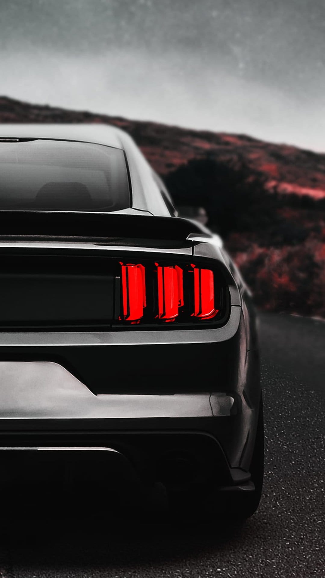 Ford Mustang triple-bar red tail lights glow on a dark asphalt road against a crimson twilight sky and hills.