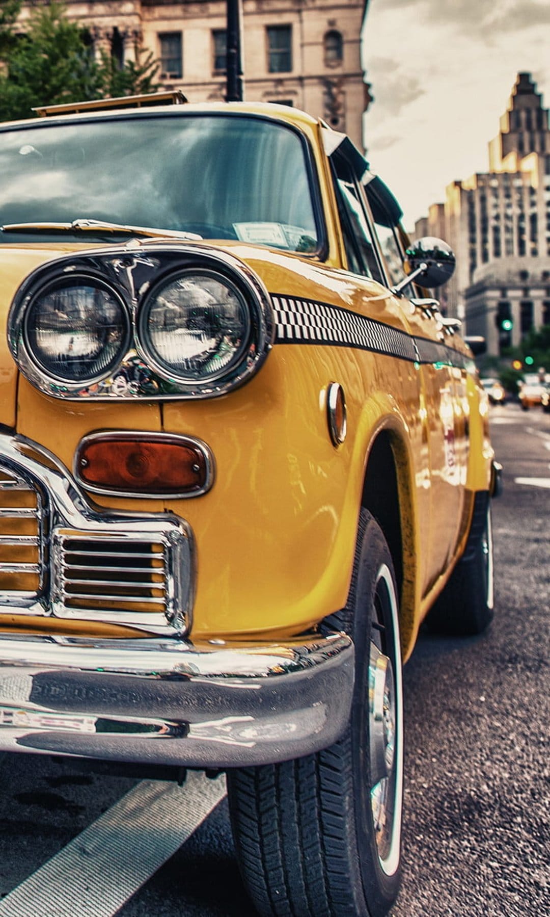 Yellow vintage taxi cab with chrome grille and double headlights parked on a blurred city street at a low angle.