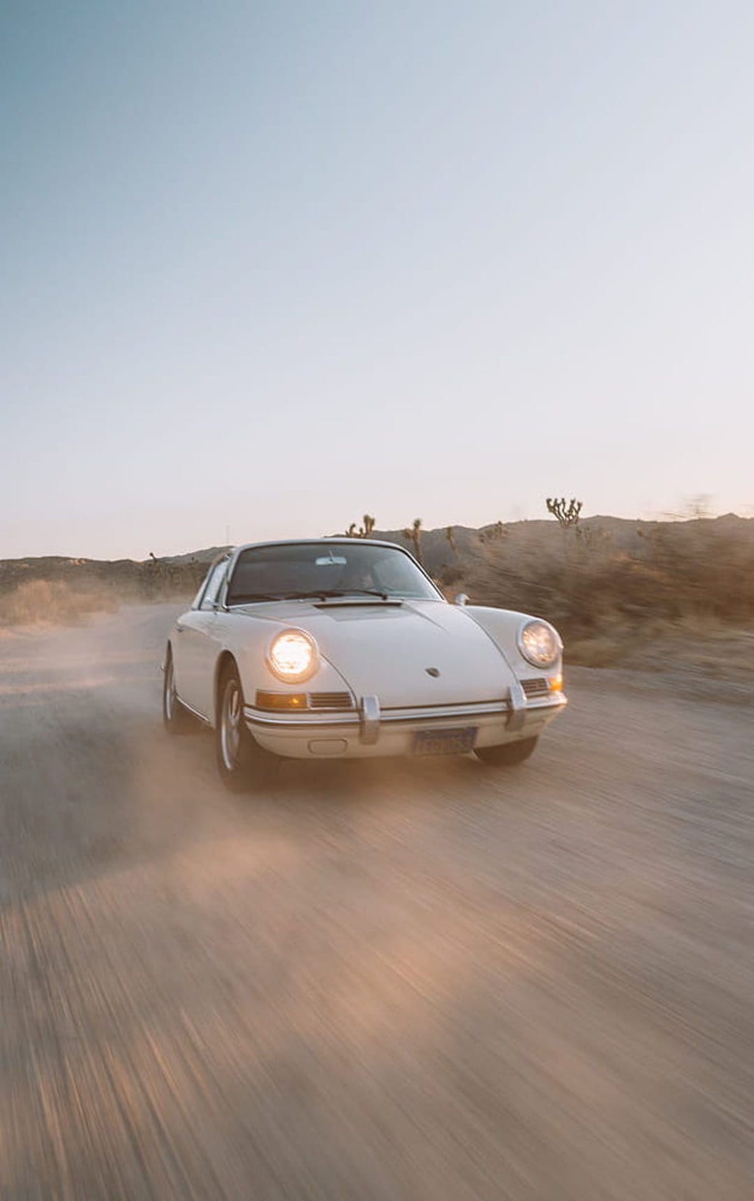 White Porsche 911 kicks up dust on a desert road with glowing headlights under a hazy golden sunset sky.
