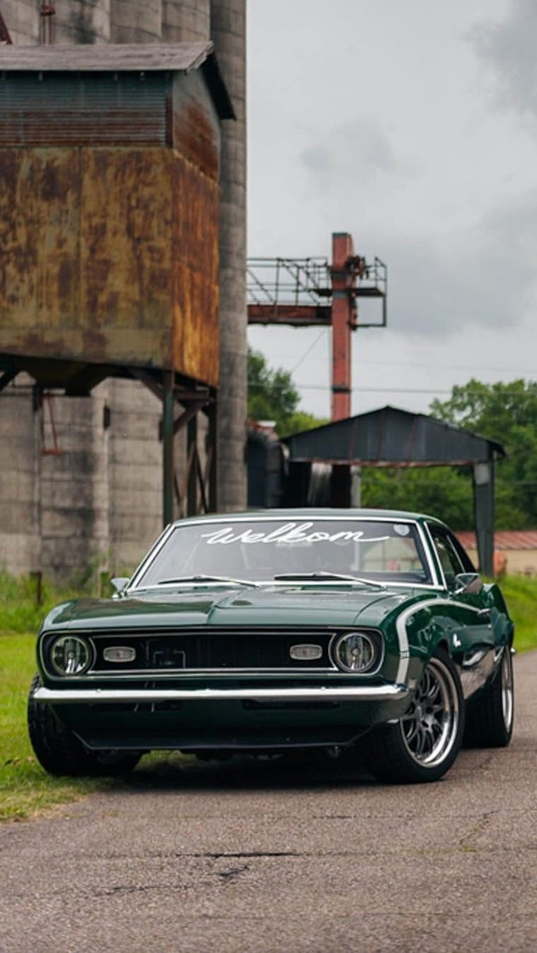 Dark green vintage Camaro parked on asphalt near weathered industrial silos with chrome accents and a windshield decal.