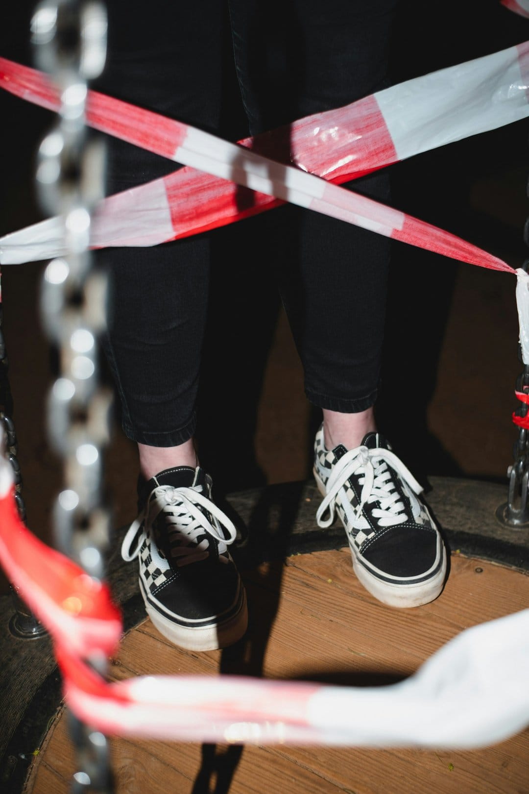 Checkerboard sneakers and black trousers stand behind a red and white caution tape with a metal chain nearby.