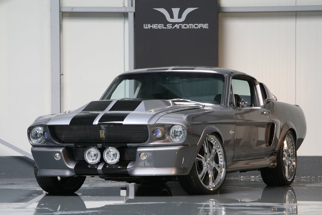 Silver Shelby GT500 Mustang with black racing stripes and polished chrome wheels in a bright studio setting.