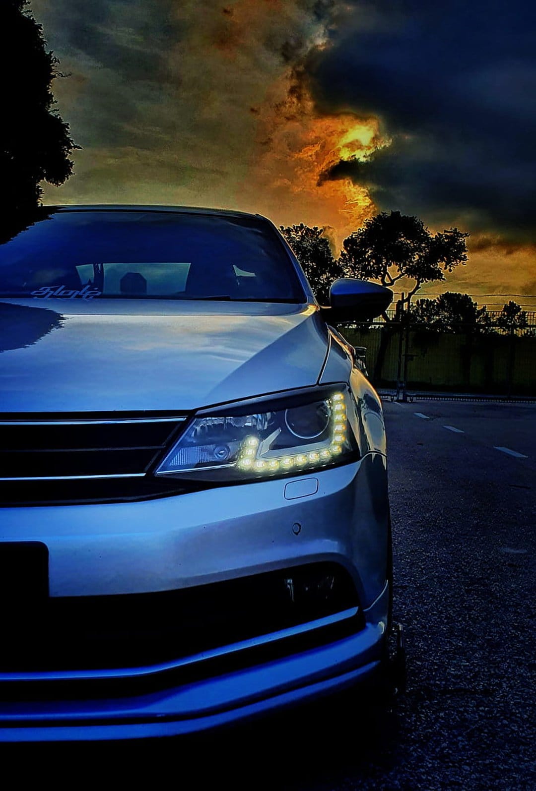 Silver Jetta LED headlights shine against a dramatic orange and grey sunset sky filled with heavy clouds.