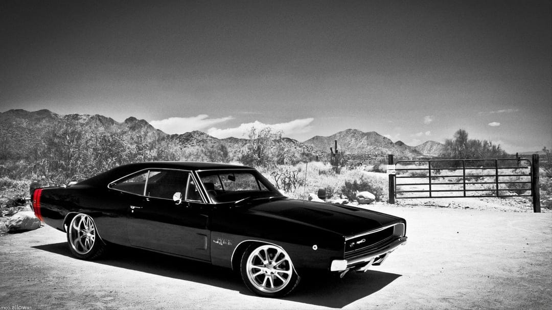 Black 1968 Dodge Charger with a red tail stripe parked in a sunny desert landscape near a metal gate.