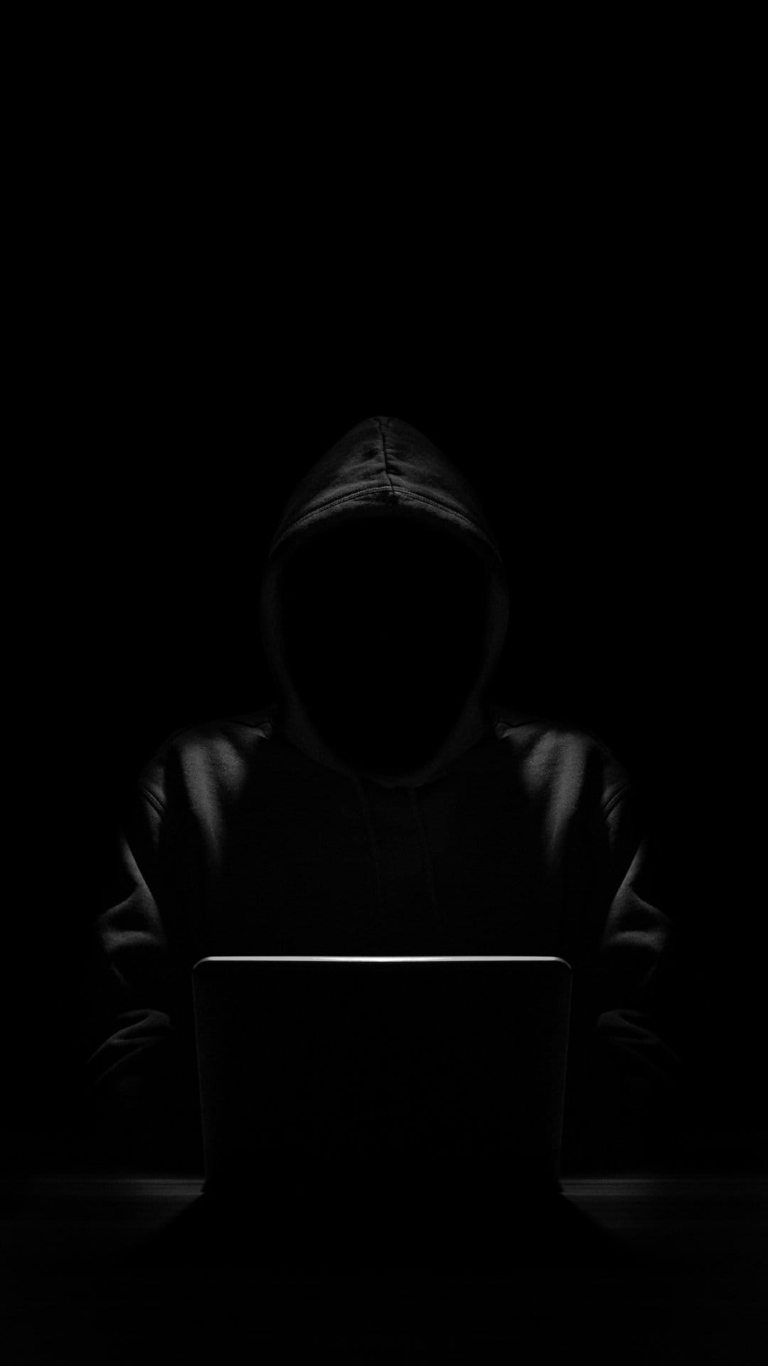 Hooded figure sitting in darkness illuminated by a glowing laptop screen with sharp blue light on their face.