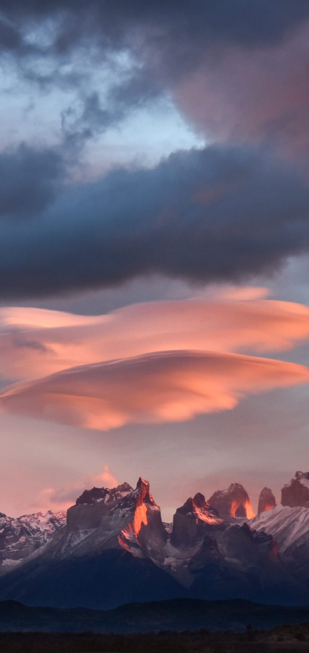 Snowy mountain peaks sit beneath glowing pink lenticular clouds and dark storm clouds during a dramatic sunset.