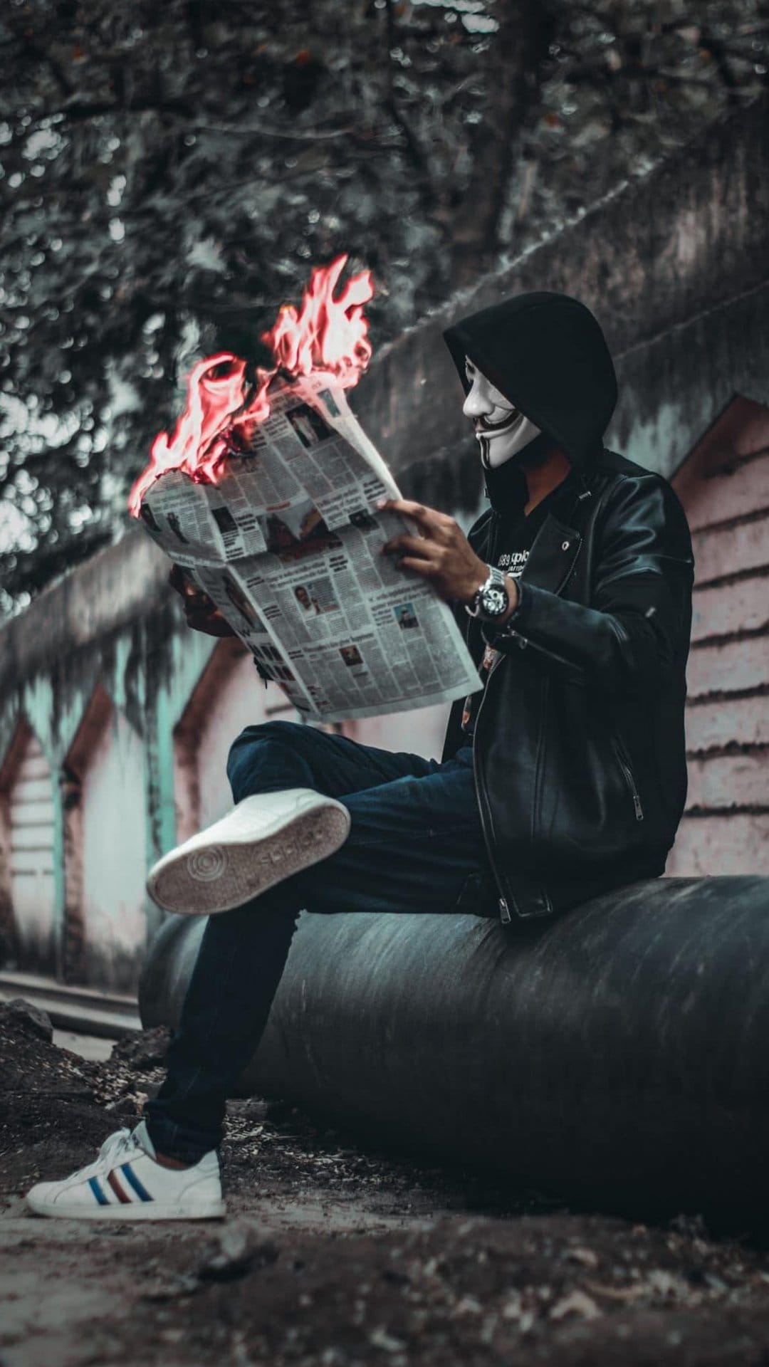 Masked rebel in a leather jacket sits on a pipe holding a burning newspaper against a blurred urban backdrop.