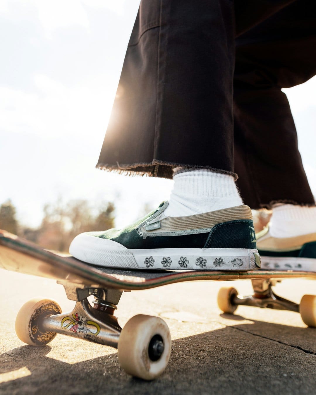 Person wearing floral Vans and dark wide-leg pants stands on a silver skateboard with yellow wheels in bright sun.