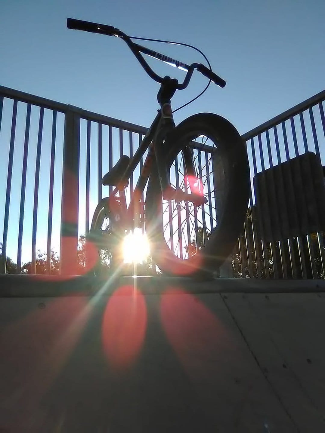 BMX bicycle silhouette against a bright sunburst and lens flare at an outdoor skatepark with metal railings.
