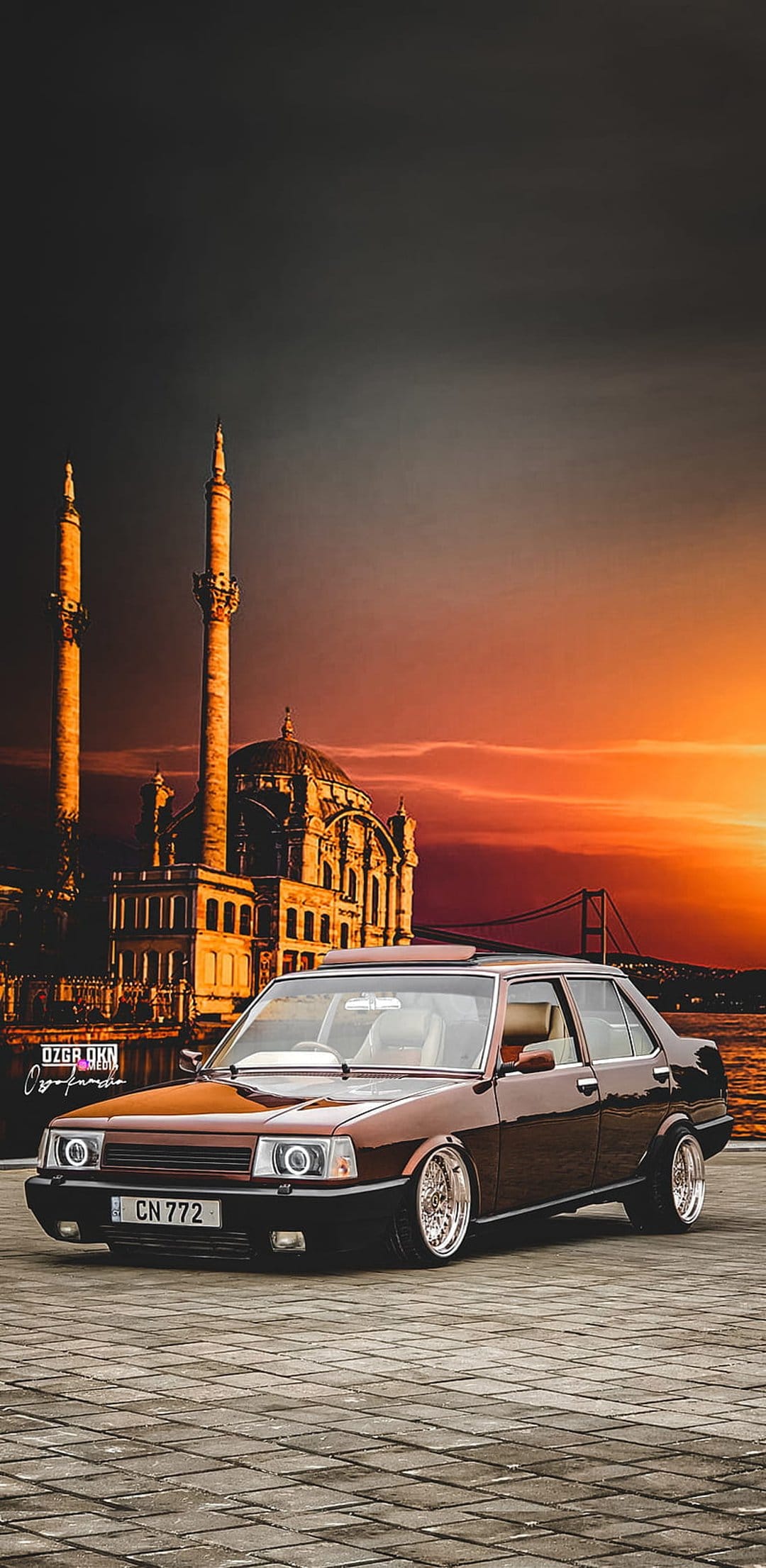Brown modified Tofaş Şahin car parked near a Turkish mosque and bridge under a vibrant orange sunset sky.