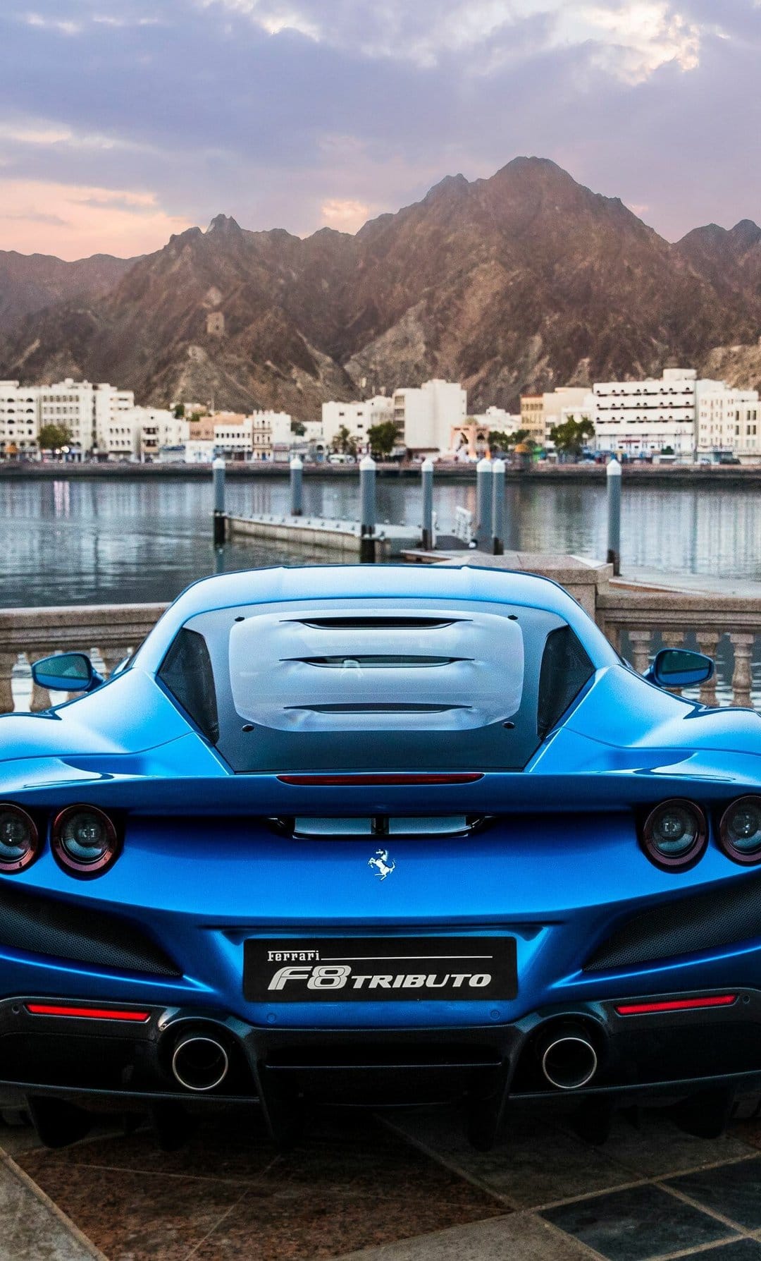 Rear view of a blue Ferrari F8 Tributo parked by a waterfront with mountains and buildings in the background.