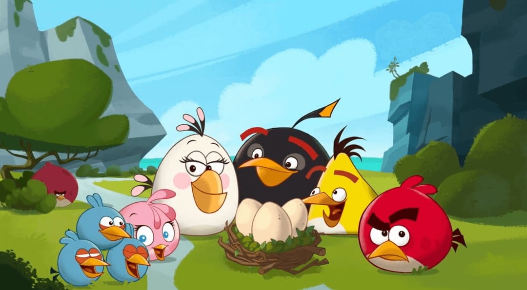 Angry Birds flock gathered around a nest of three eggs in a sunny green field with blue skies and rocky cliffs.