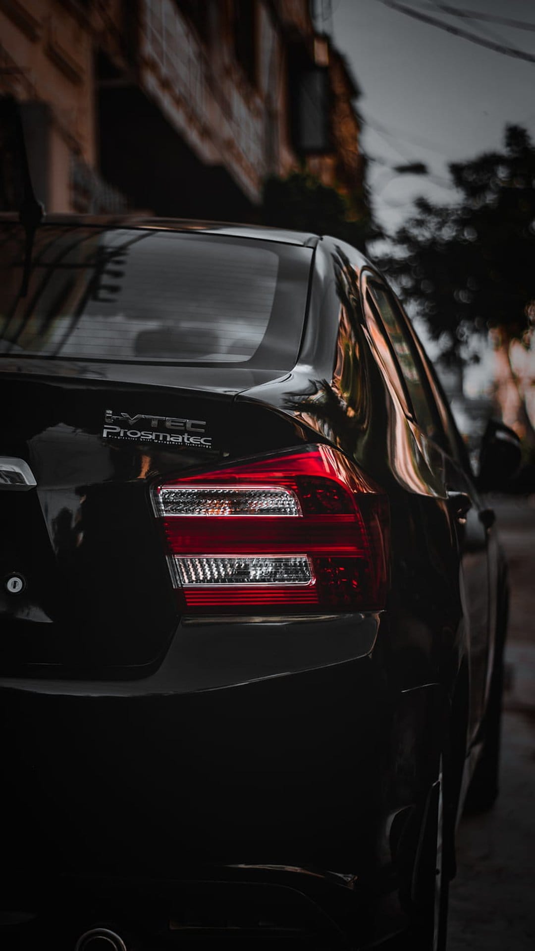 Honda City sedan rear taillight and chrome badging on a polished black surface with urban bokeh background.
