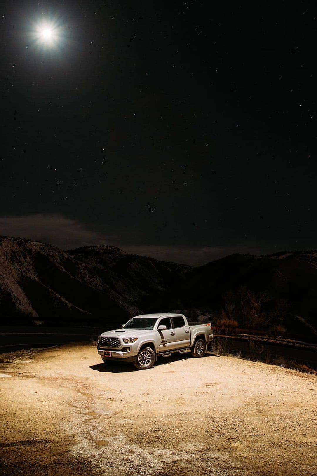 Silver Toyota Tacoma pickup truck parked on a dirt lot under a bright moon, stars, and silhouetted mountains.
