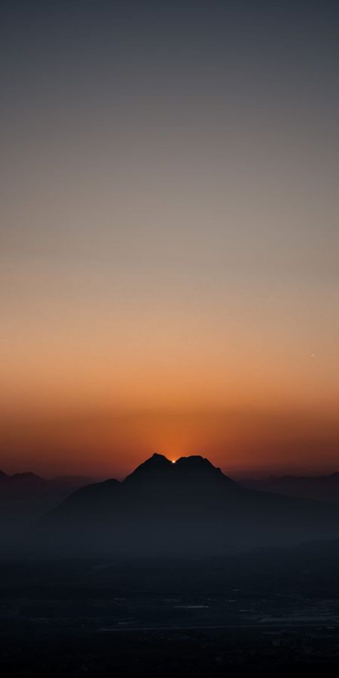 Mountain peaks silhouette against a gradient orange and grey sky as the sun dips behind a sharp rocky ridge.