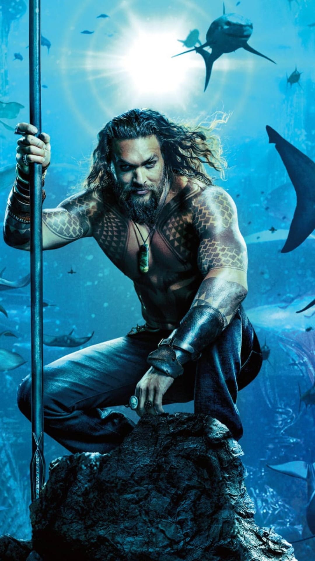 Jason Momoa as Aquaman holds a trident on a rock with sharks and sunlight filtering through blue water.
