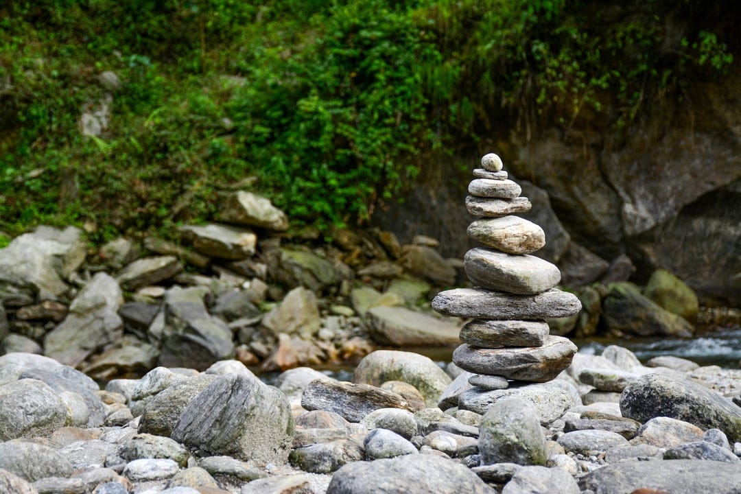 Balanced grey stone cairn on a riverbank beside a rushing stream surrounded by vibrant green forest plants.
