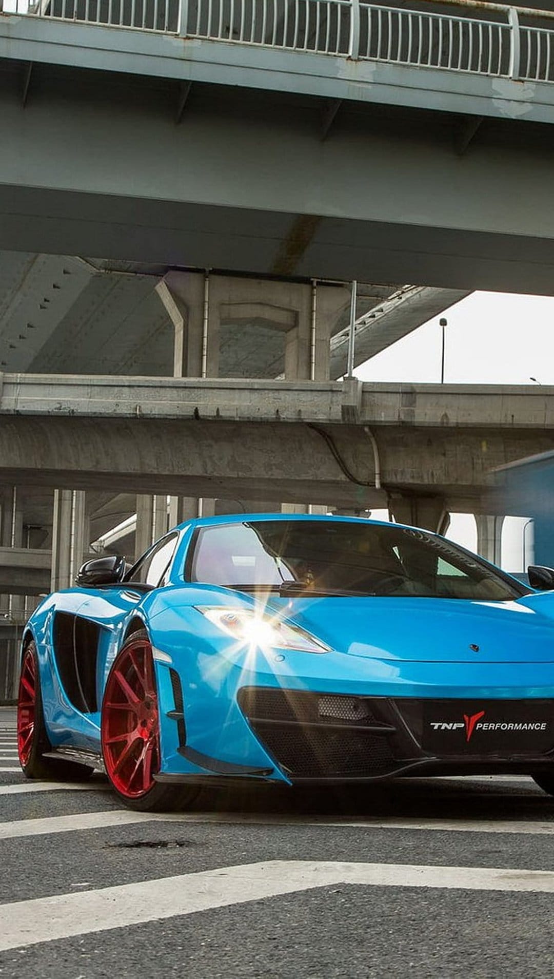 Blue McLaren supercar with red wheels parked on a crosswalk under a concrete bridge with sunlight on headlights.