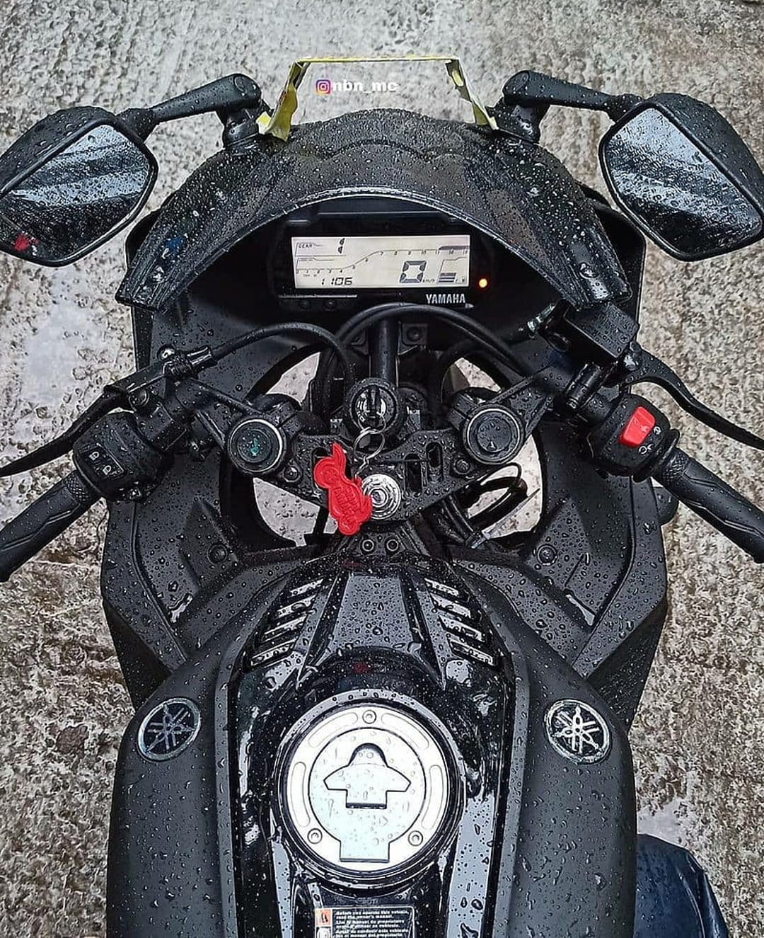Black Yamaha R15 motorcycle with water droplets on the fairing and digital dashboard during a rainy day.