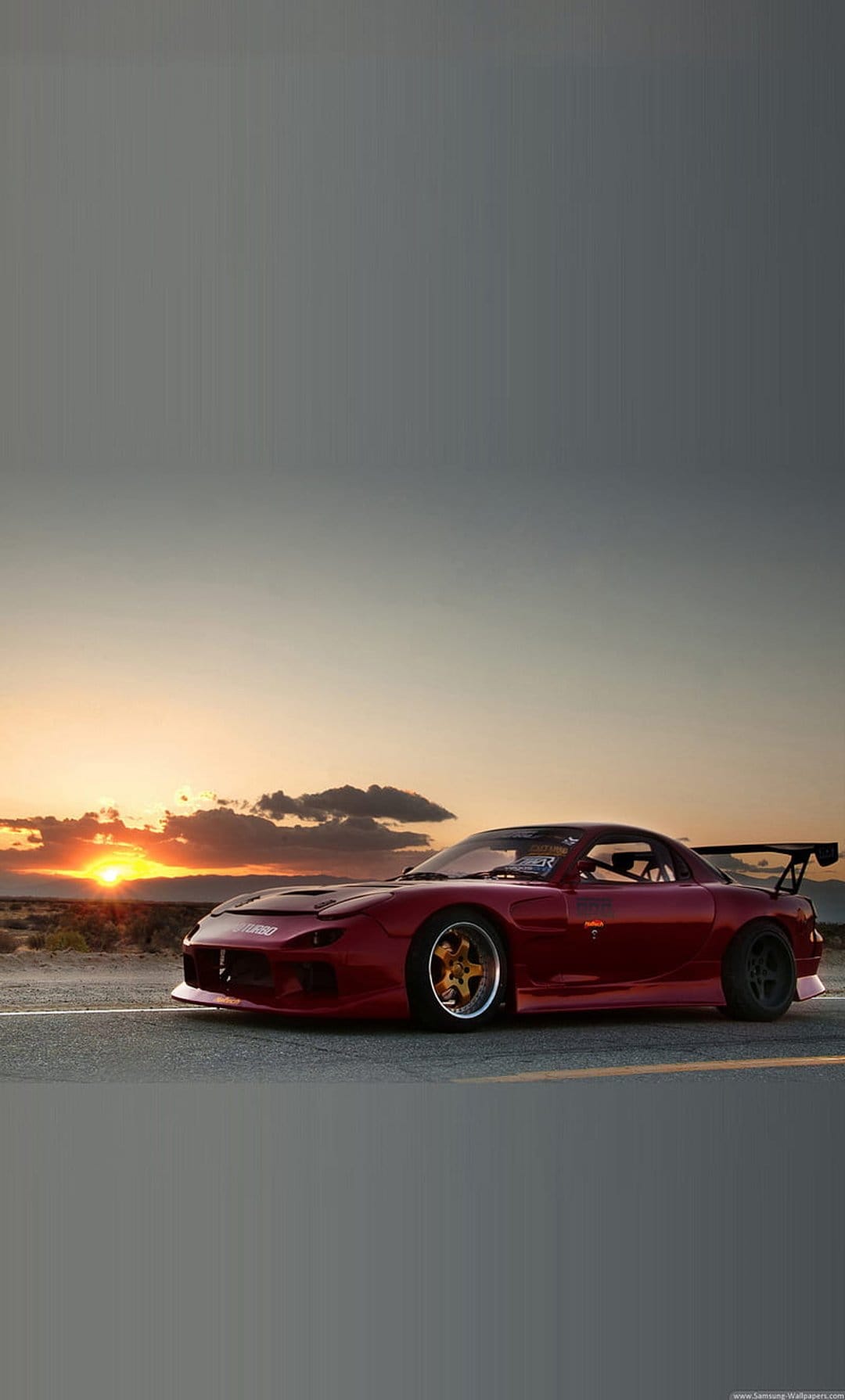 Red Mazda RX-7 sports car with a large spoiler parked on a desert road during a vibrant orange sunset.