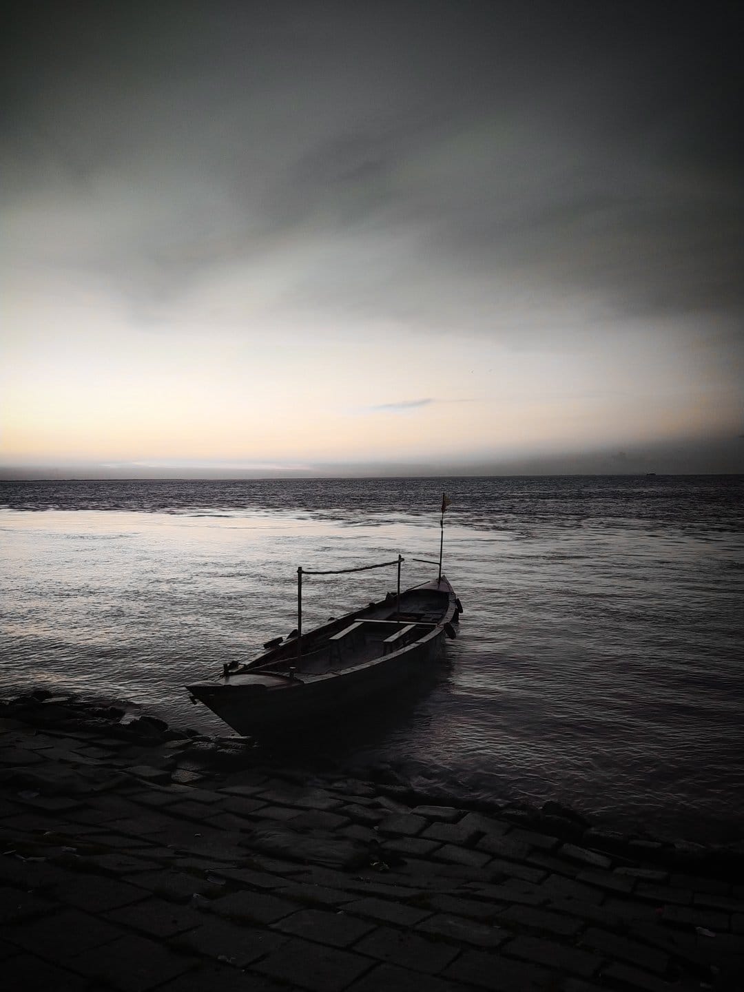 Wooden boat drifts on dark rippling water near a rocky shore under a dim gray twilight sky at the edge of a lake.