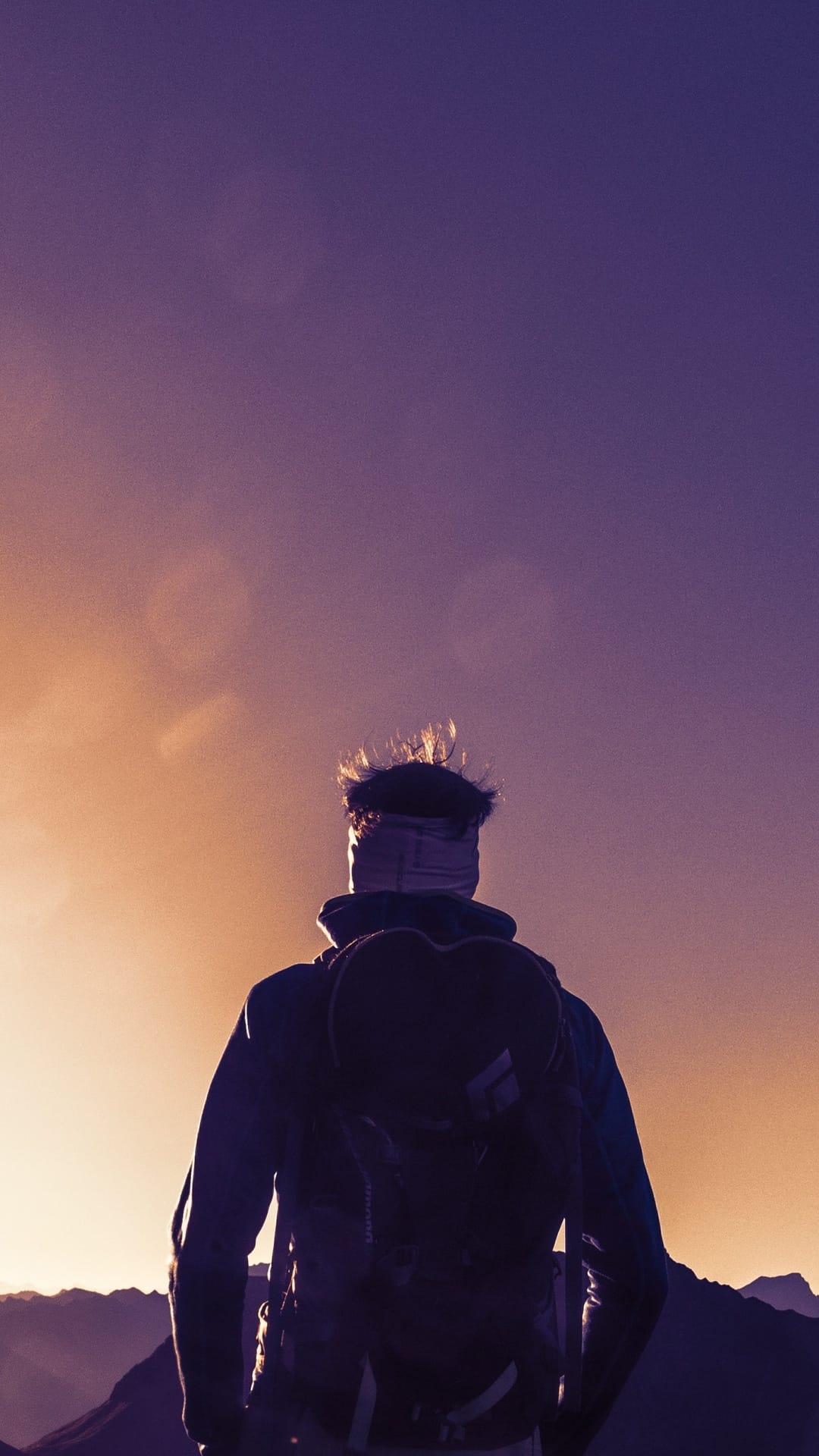 Hiker with a backpack stands on a rocky summit silhouetted against a purple and orange sunset gradient sky.