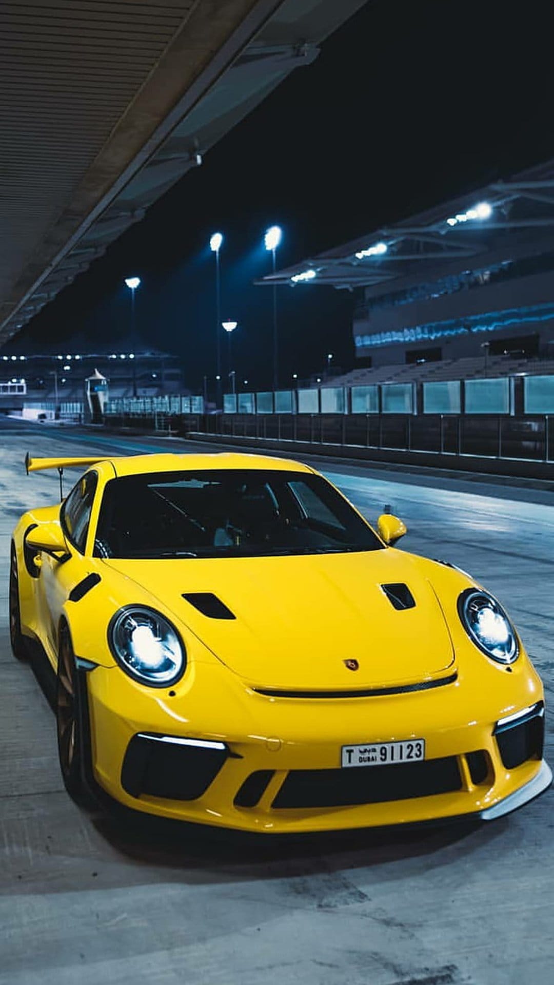 Yellow Porsche 911 GT3 RS with glowing headlights parked on a dark racetrack at night under stadium floodlights.