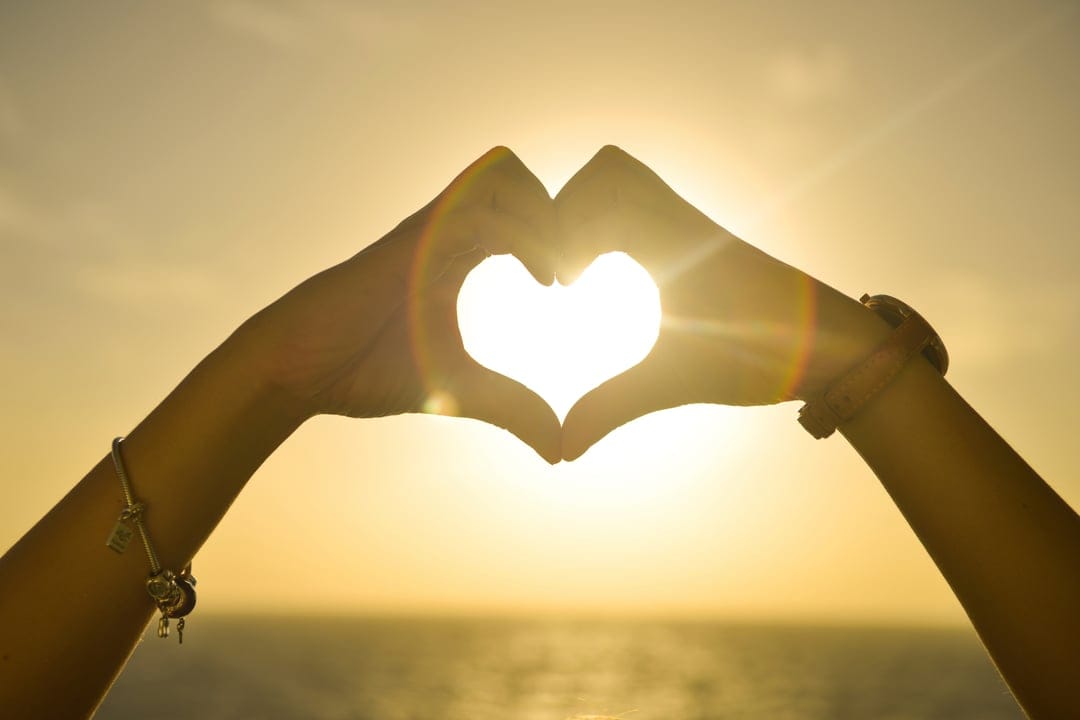 Silhouetted hands forming a heart shape with sunbeams glowing through the center over a calm ocean.