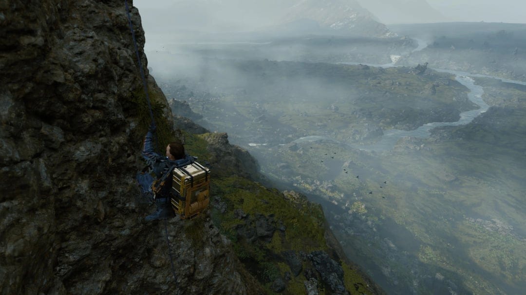 Sam Porter Bridges climbs down a rocky cliff face with a blue rope overlooking a vast, misty river valley.