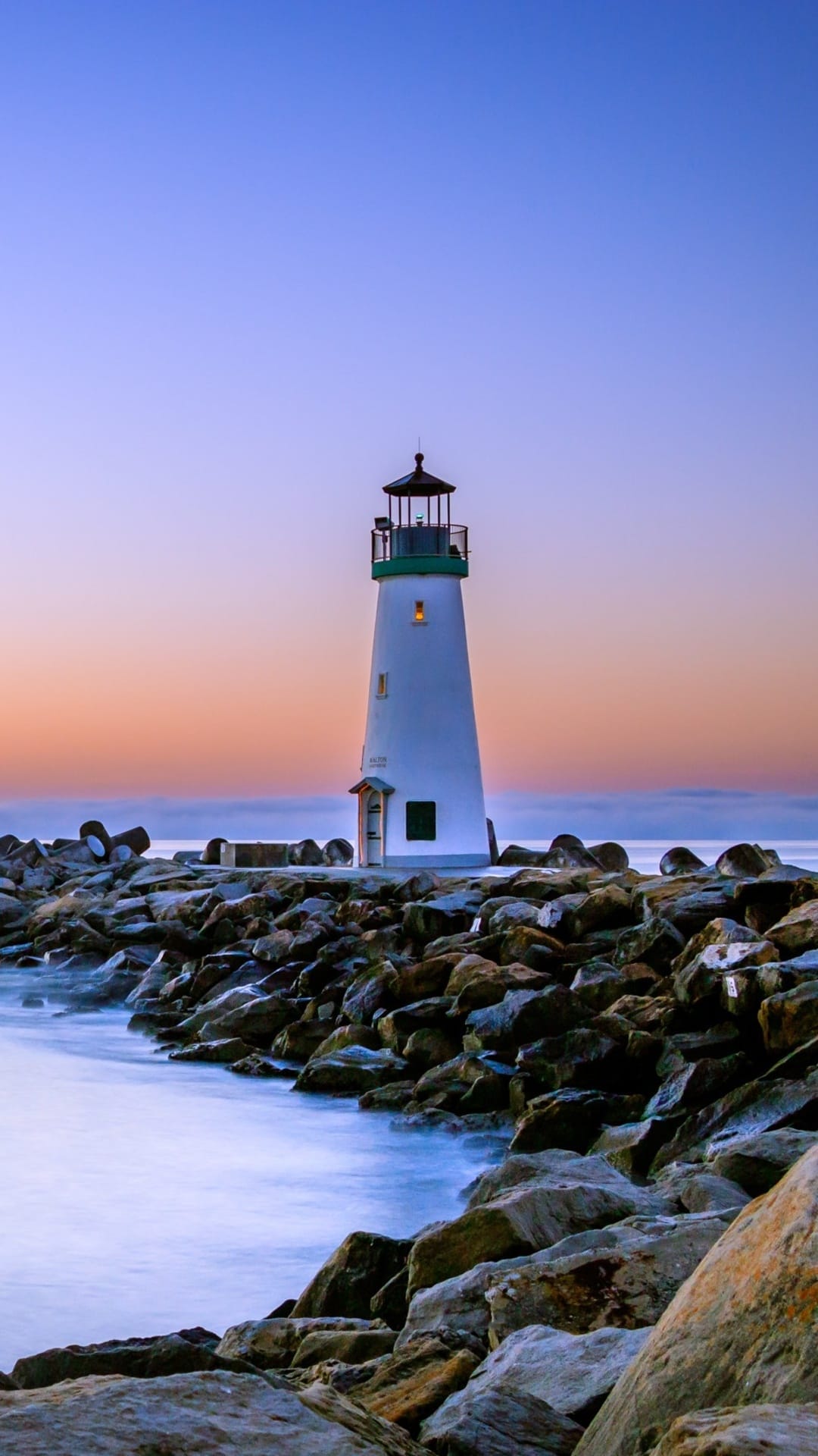 White lighthouse with a green cap stands on a rocky breakwater during a soft orange and blue sunset.