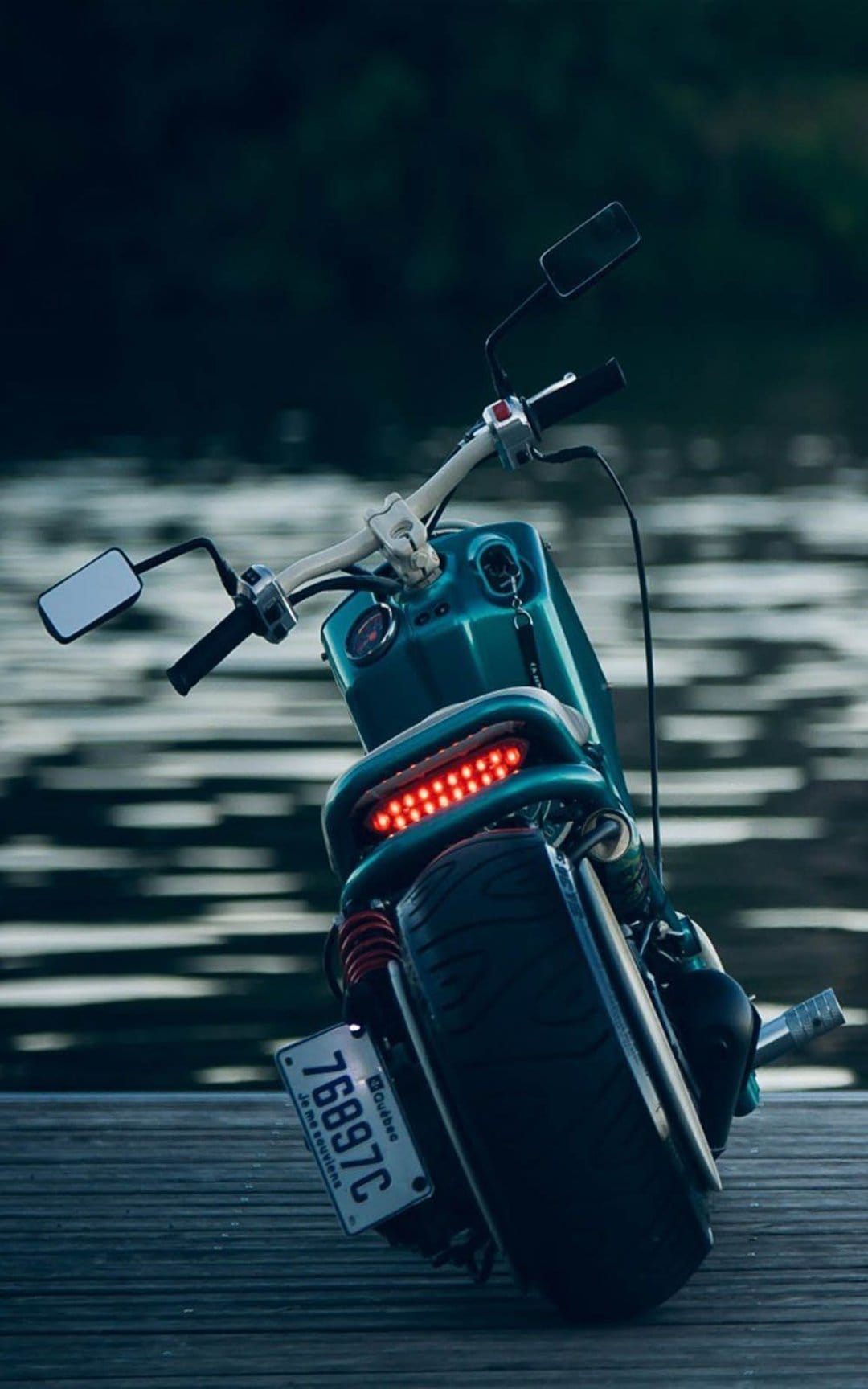 Teal scooter with a red LED taillight and wide tire on a weathered wooden dock over dark rippling water.