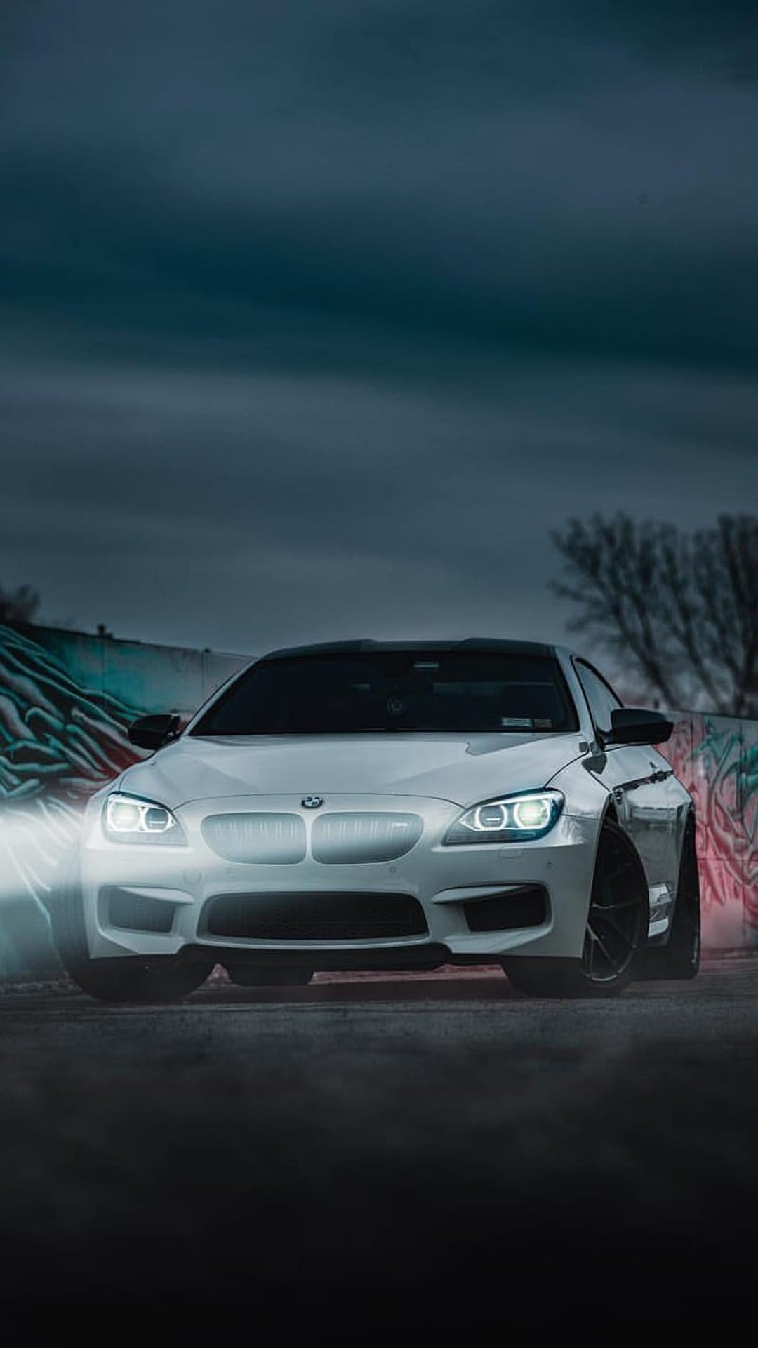 White BMW M6 coupe with lit headlights parked before a colorful graffiti wall at night with drifting mist.