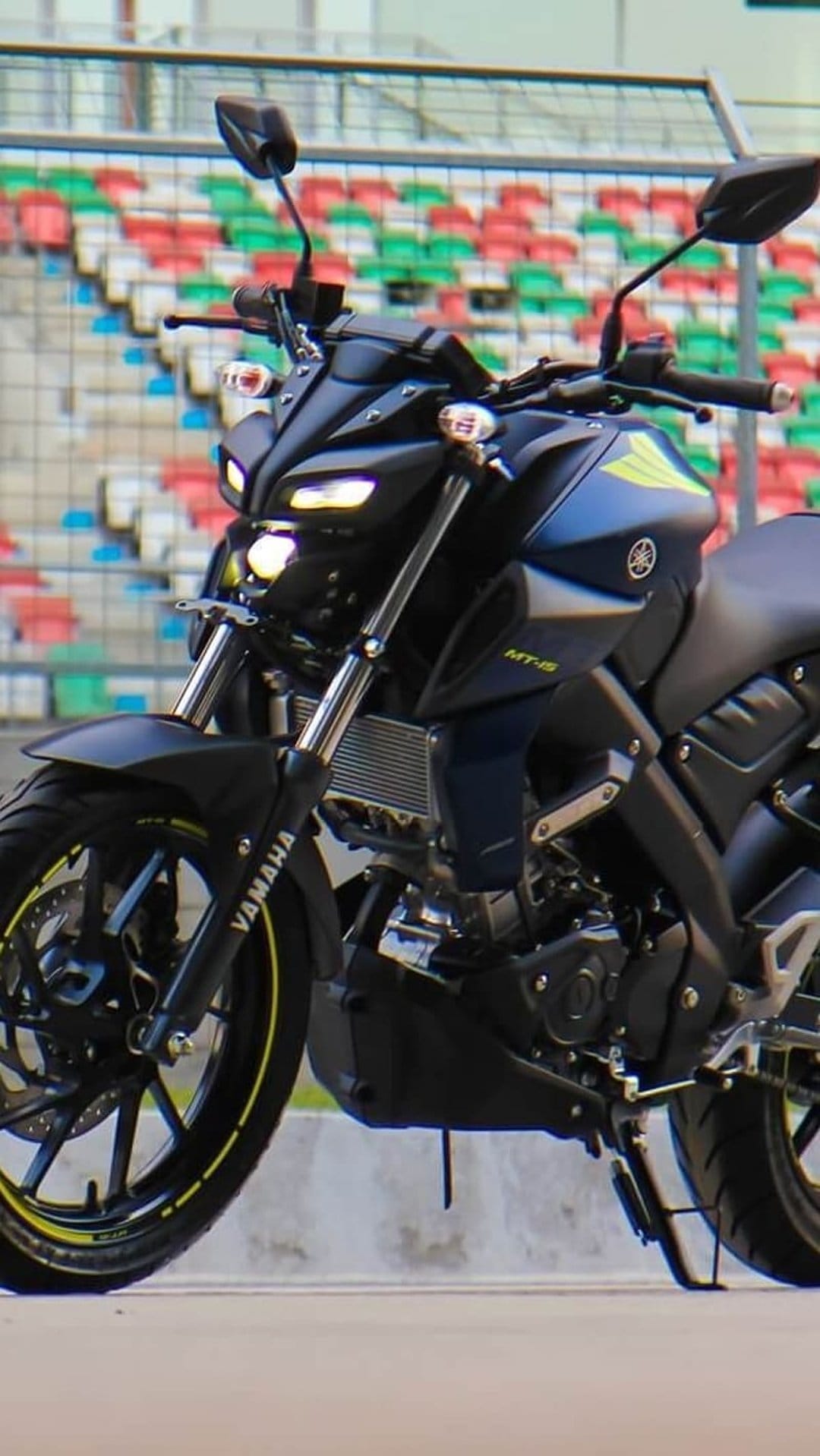Yamaha MT-15 motorcycle with yellow wheel accents parked in front of blurred red and green stadium seating rows.
