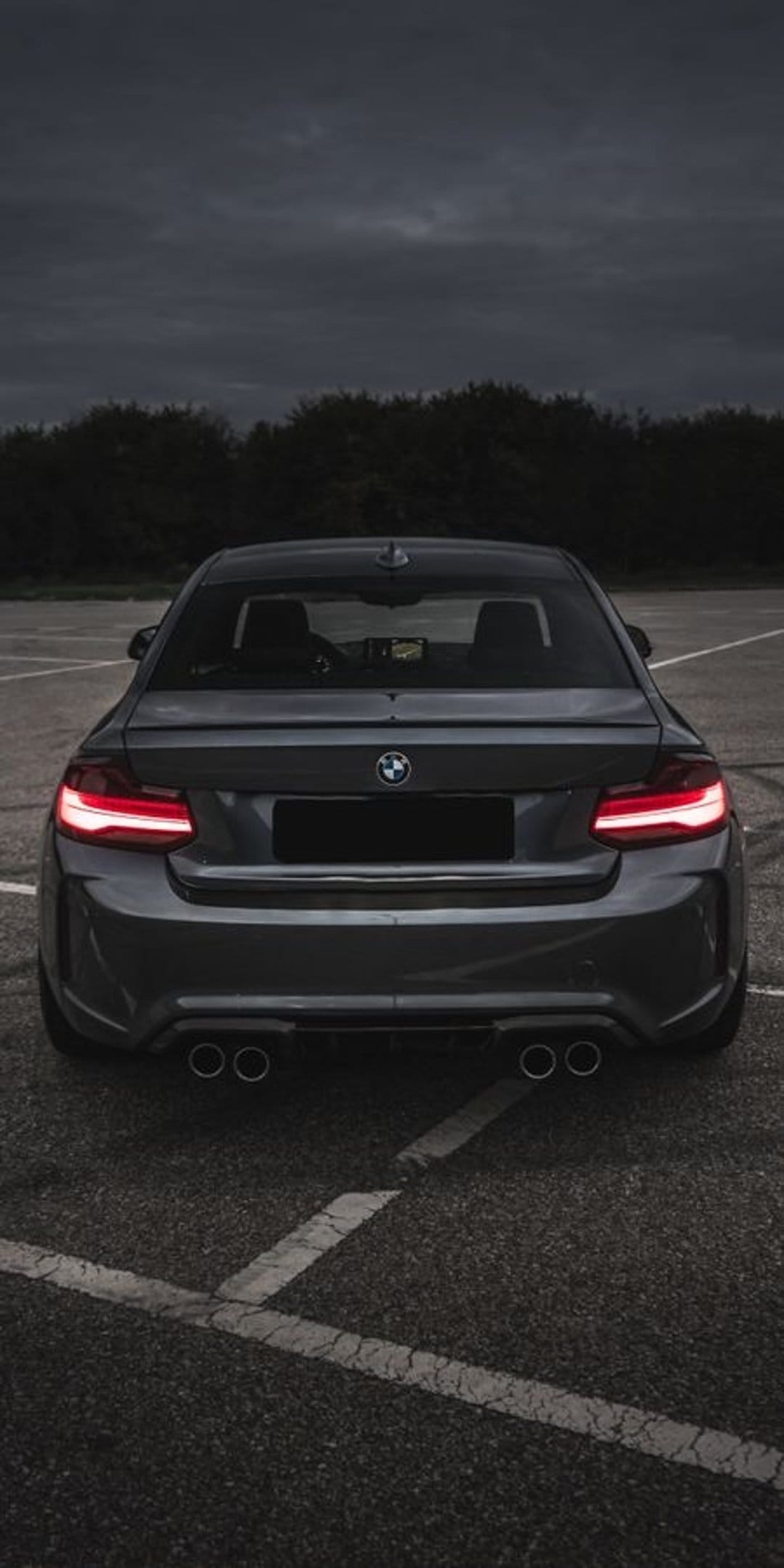 BMW M2 Coupe rear view showing glowing red LED taillights and quad exhausts on dark asphalt under a moody sky.