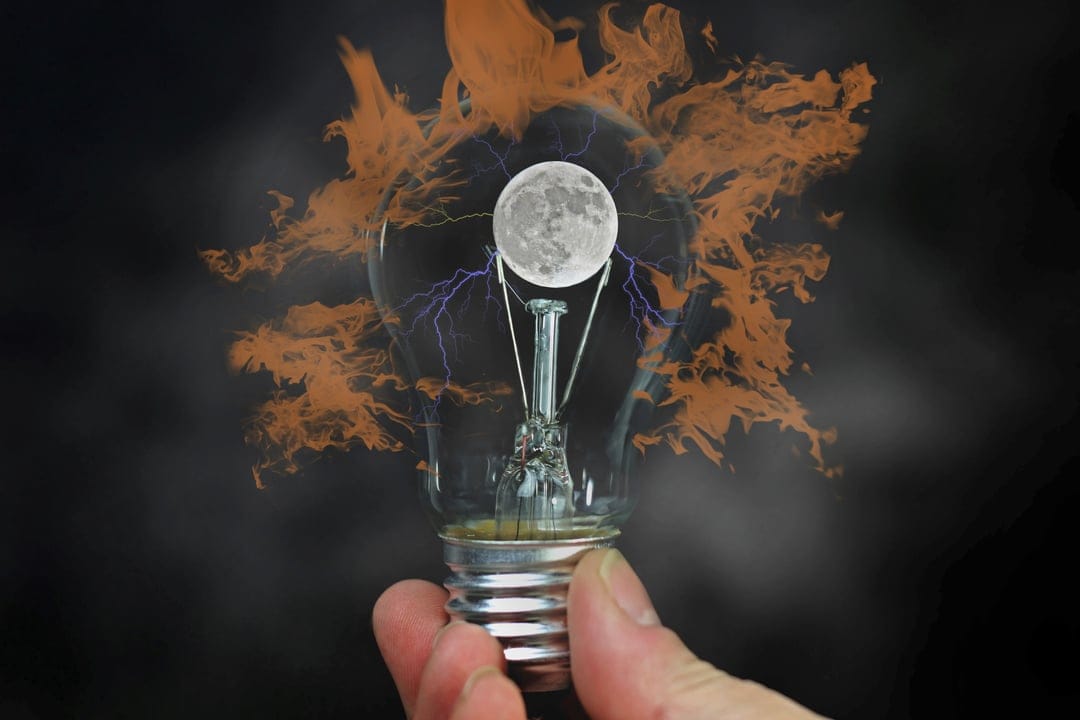 Hand holds a glass bulb containing a full moon with purple lightning amidst swirling orange fire and smoke.