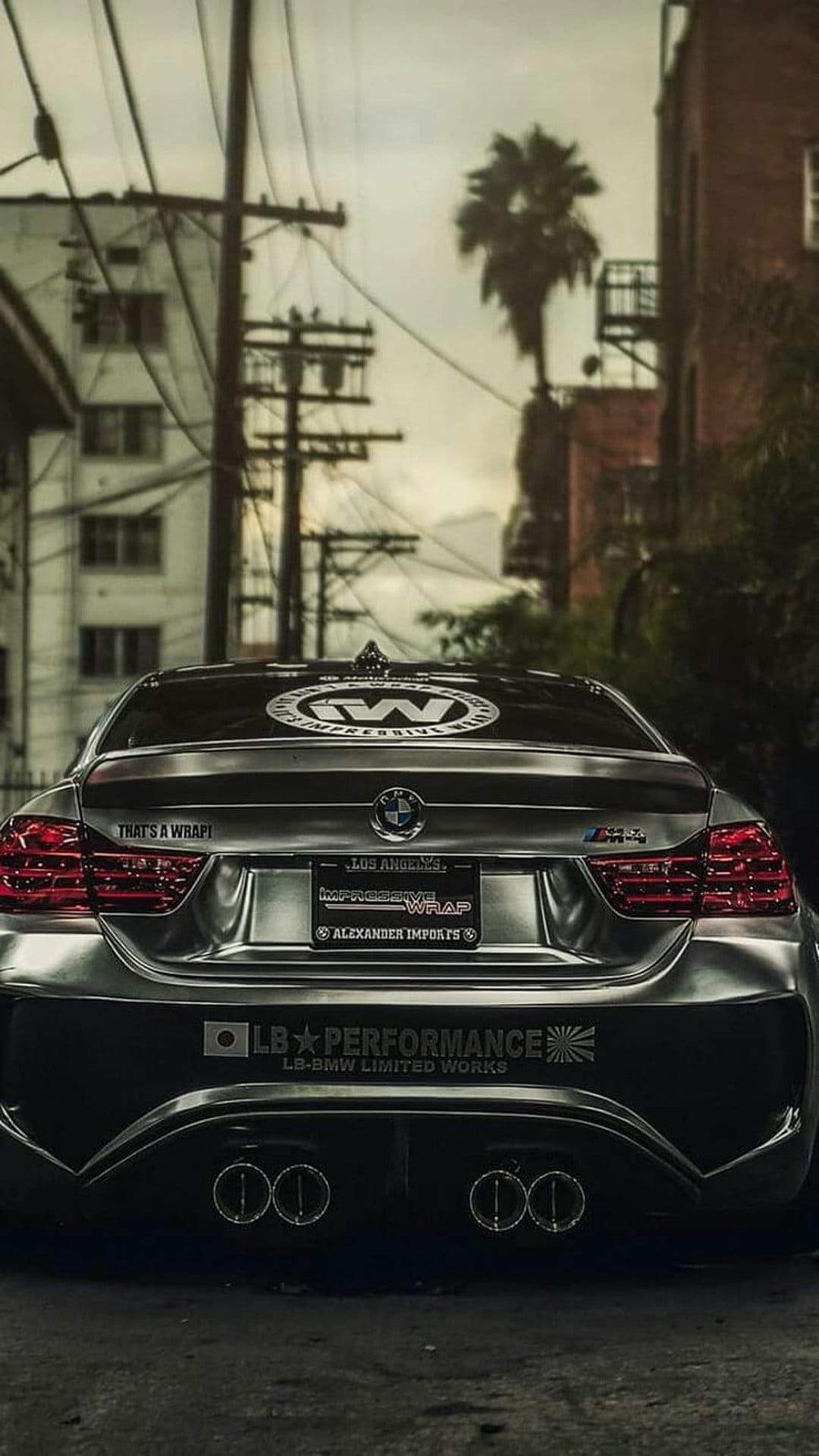 Silver satin BMW M4 with a widebody kit parked in an urban alley beneath power lines and a distant palm tree.