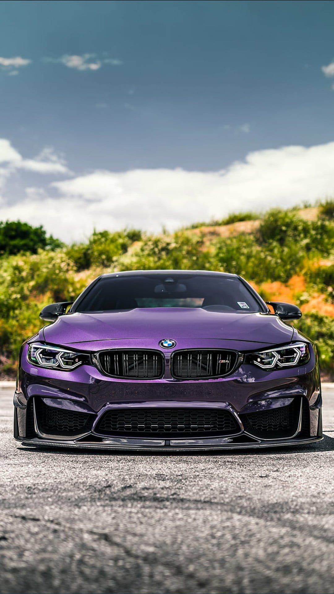 Purple BMW M4 coupe parked on asphalt with lush green foliage and a blue cloudy sky in the background.