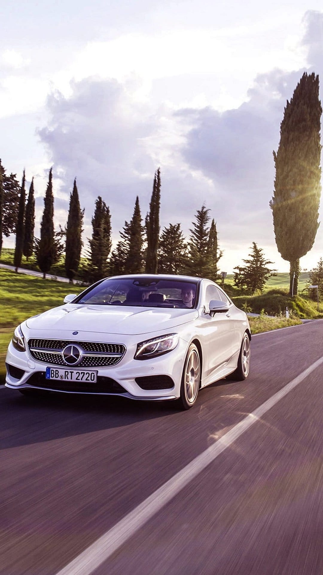 White Mercedes S-Class Coupe drives along a winding road lined with tall cypress trees under a dramatic cloudy sky.