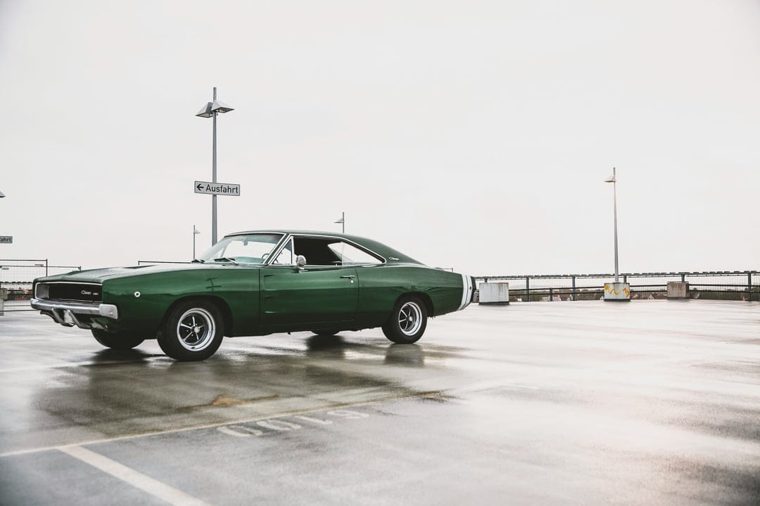 Classic green Dodge Charger on rain-slicked pavement with chrome trim and a moody urban parking lot backdrop.