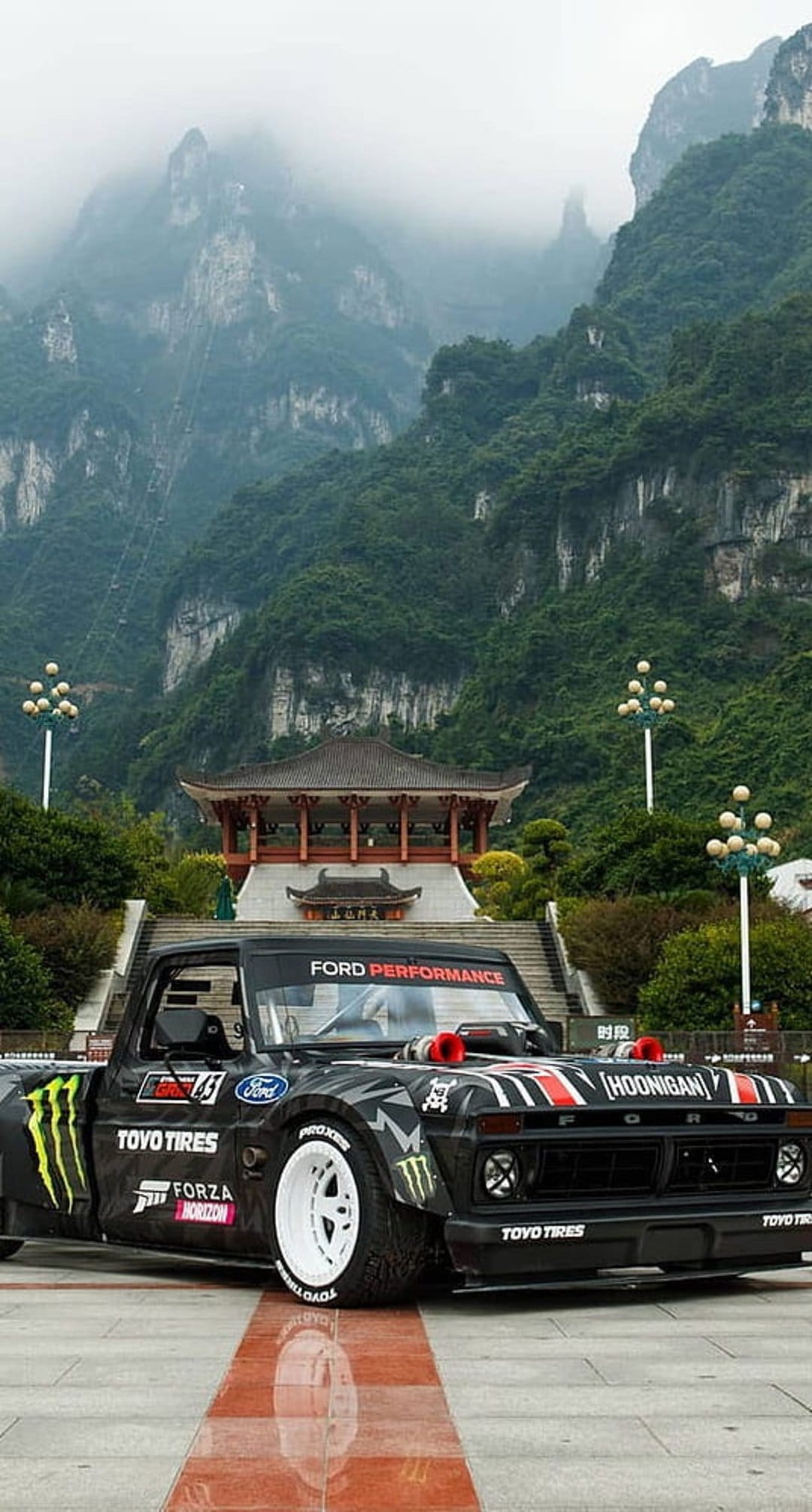 Black Ford pickup with Monster Energy decals parked near a traditional temple amid misty green mountains.