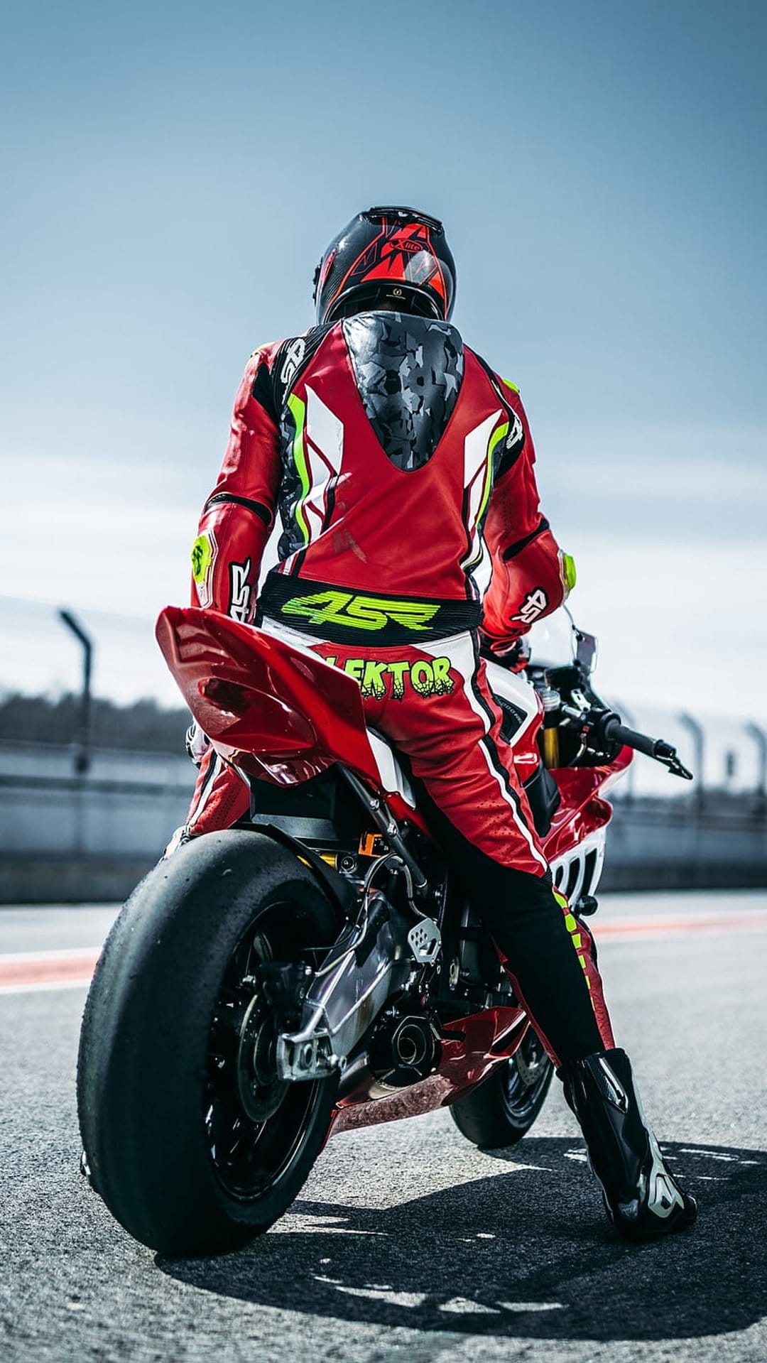 Motorcyclist in a red and white racing suit sits on a sportbike on a sunlit track with sharp asphalt details.