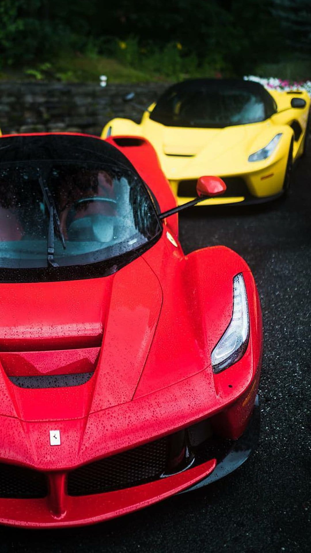 Red Ferrari with water droplets on the hood parked near a yellow car on wet asphalt during a rainy day.
