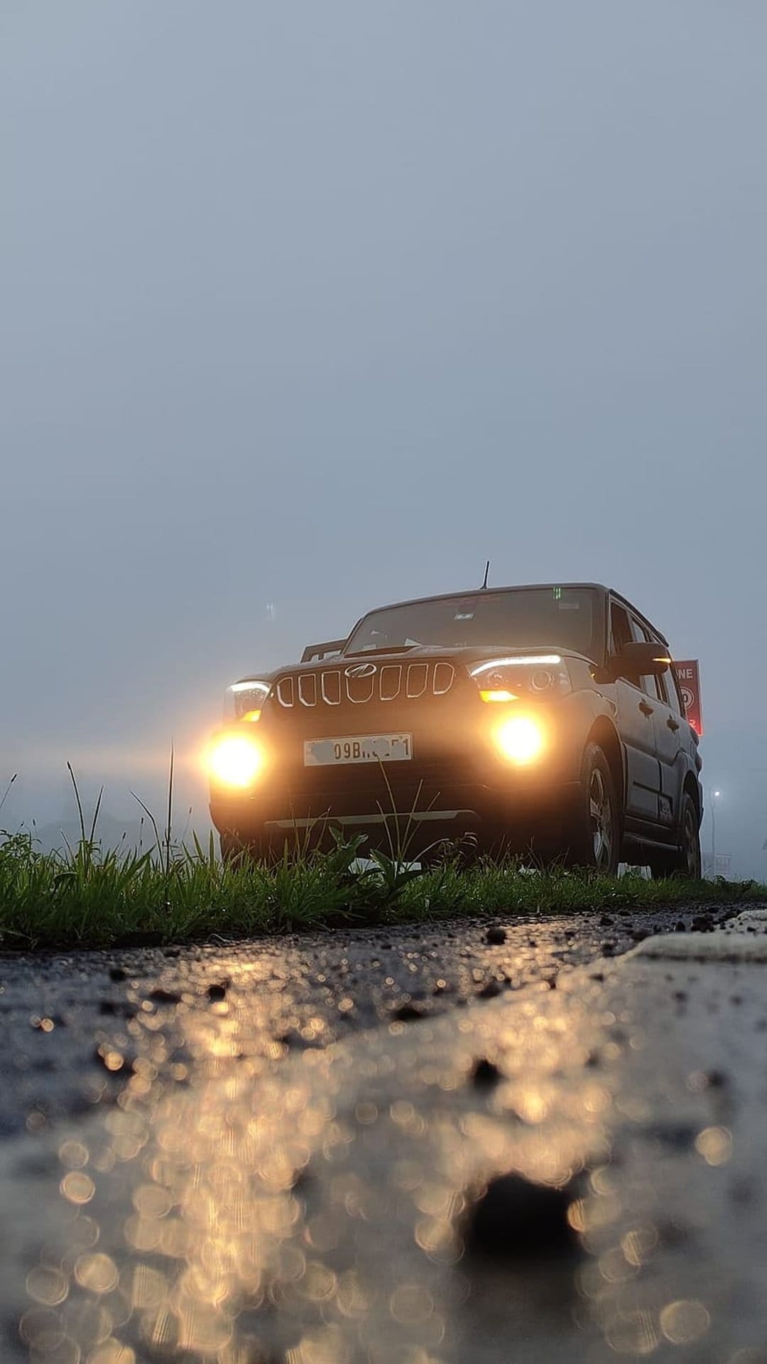 Mahindra Scorpio SUV with glowing headlights on a wet asphalt road surrounded by thick gray fog and green grass.