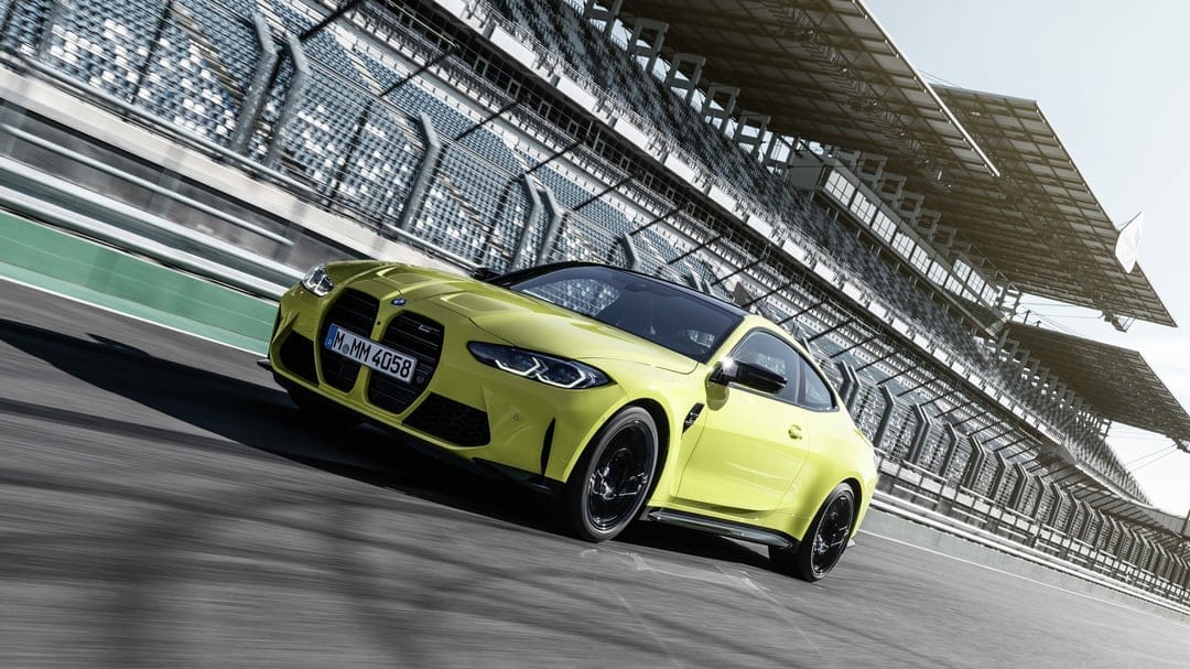 Yellow BMW M4 Coupe speeding on a race track with motion blur and large stadium grandstands in the background.