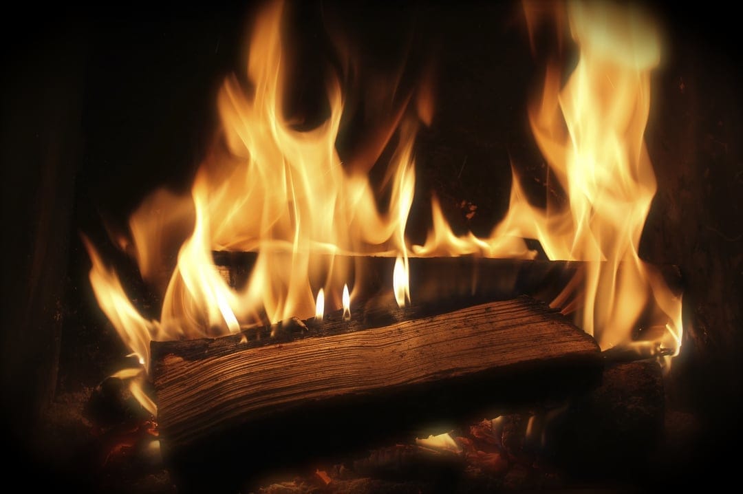 Glowing orange fire engulfing a large charred log inside a dark fireplace with flickering embers and sparks.